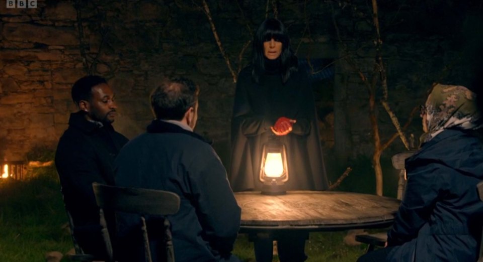 A scene from The Traitors showing four contestants seated around a table at night, listening to a woman in a black cloak.