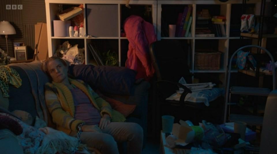 Screenshot of a messy room, a woman chained to a couch, and a person in a pink jacket in the background.