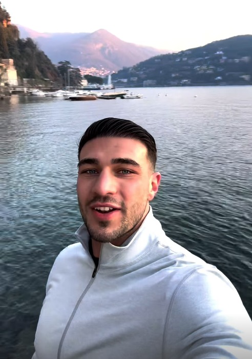 Man in light gray zip-up jacket by a lake with mountains in the background.